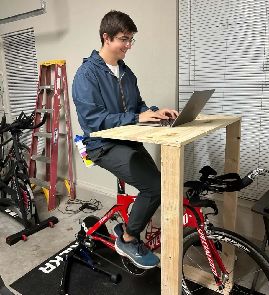 Alex riding a trainer in a garage, laptop on a plywood standing desk built over the handlebars